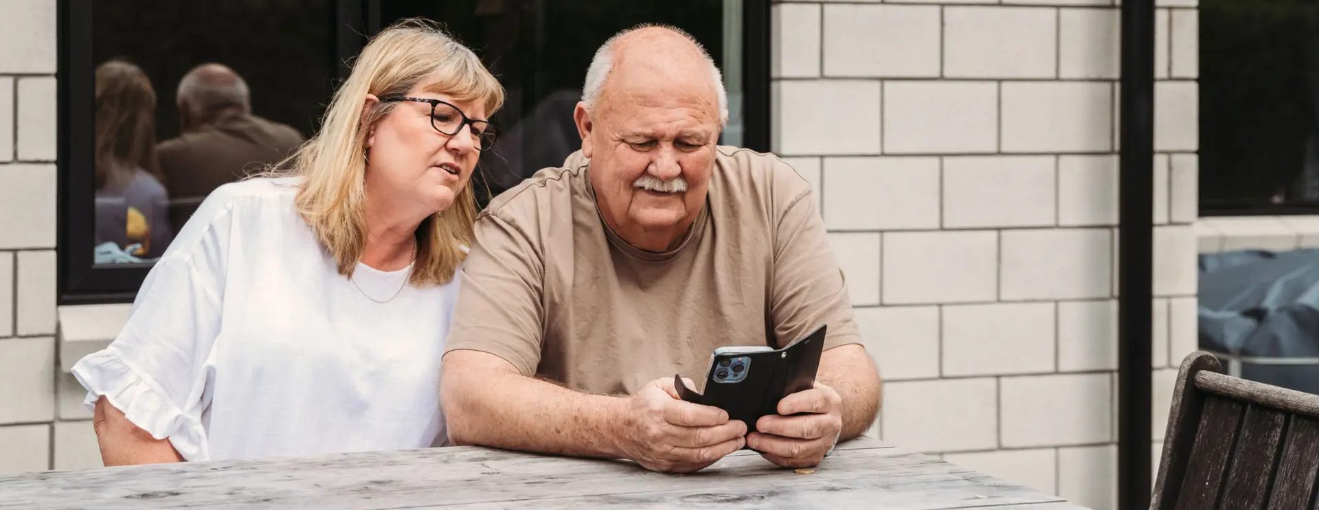 An older couple sitting at an outdoor table, looking at a smartphone together.