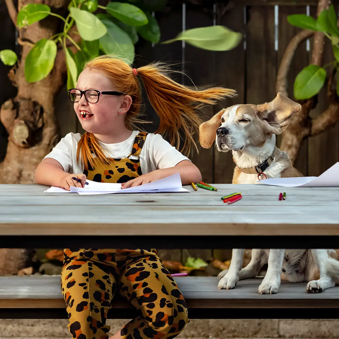 Child drawing at an outdoor table with a dog sitting beside them.