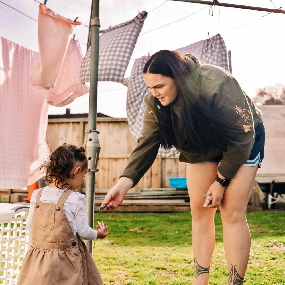 A woman bends down in a backyard to hand an item to a young child near a clothesline.