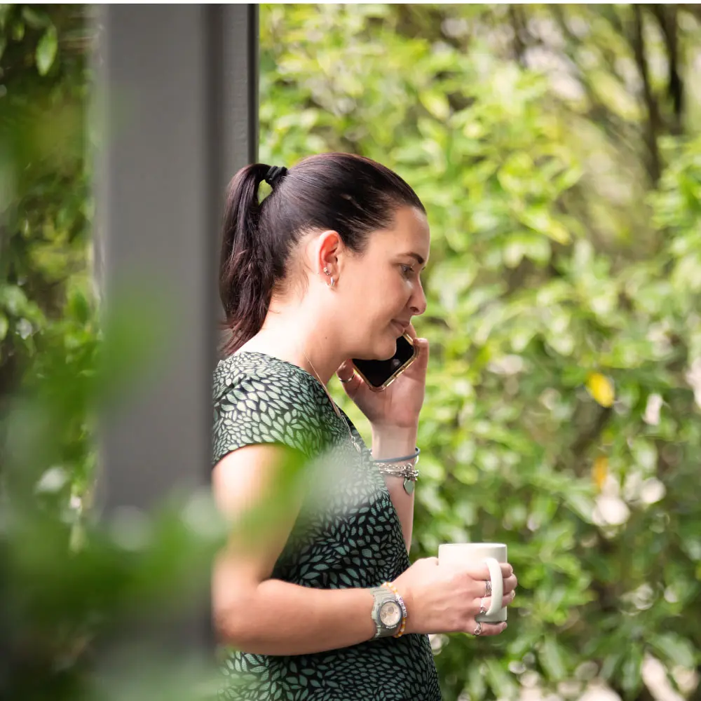 A woman stands outdoors talking on a mobile phone, with green trees and foliage in the background.