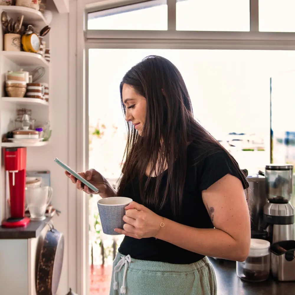 Woman standing in her kitchen, looking a her phone while holding a mug