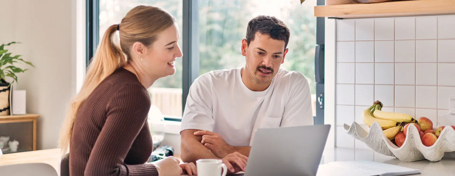 Two people standing in a kitchen, looking at a laptop together on a bench.