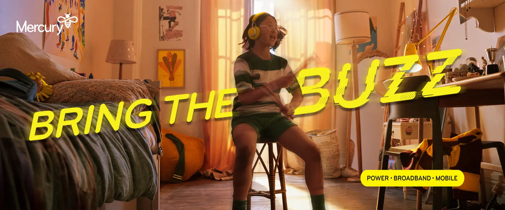 Young person wearing headphones and playing drumsticks while sitting in a bedroom.