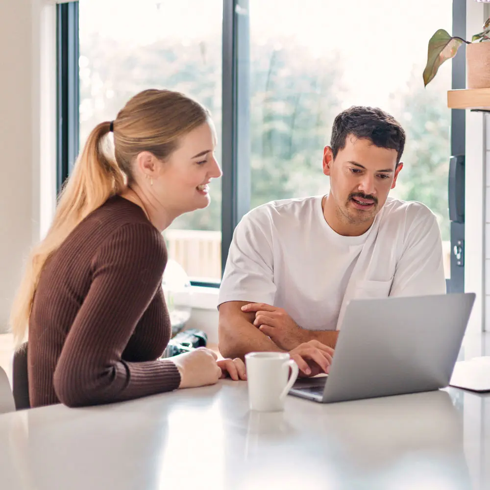 Two people standing in a kitchen, looking at a laptop together on a bench.