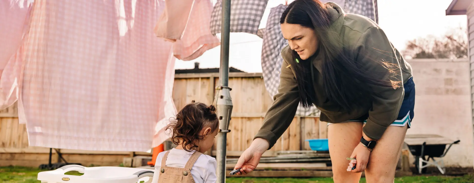 A woman bends down in a backyard to hand an item to a young child near a clothesline.