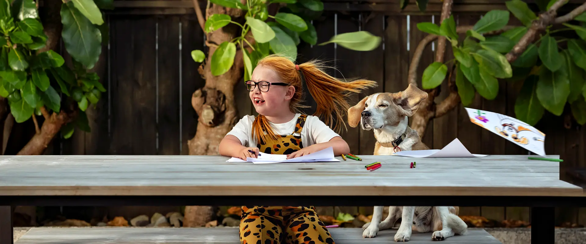 Child drawing at an outdoor table with a dog sitting beside them.