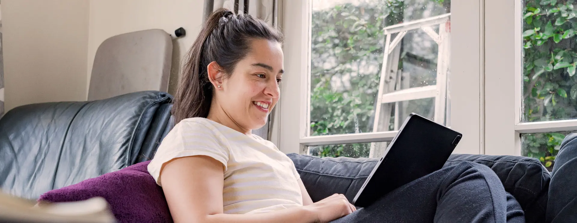 A woman sitting on a couch at home, smiling while using a tablet.