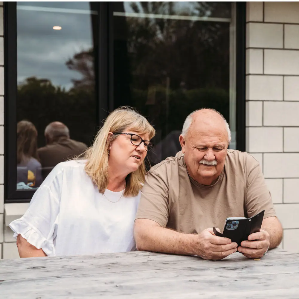 An older couple sitting at an outdoor table, looking at a smartphone together.