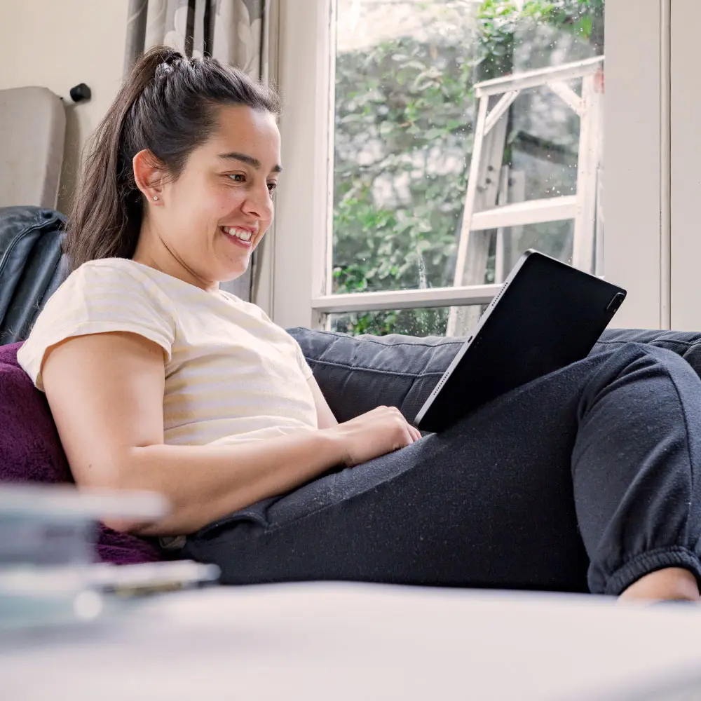 A woman sitting on a couch at home, smiling while using a tablet.